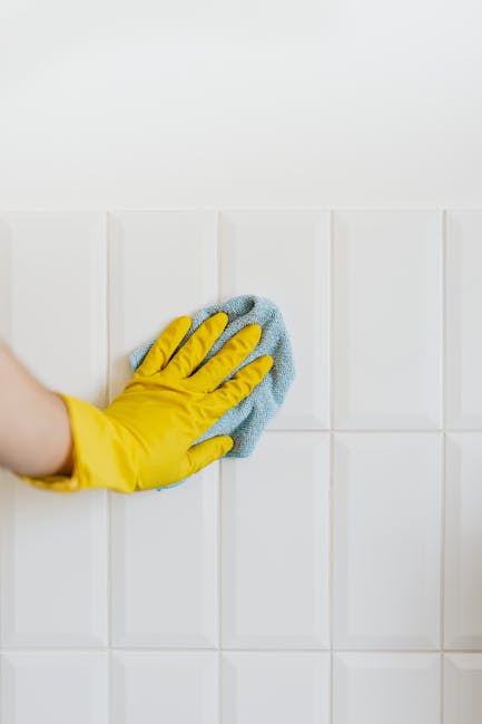 A person wearing a yellow rubber glove is using a blue cleaning cloth to wipe white ceramic tiles on a wall in a kitchen or bathroom. The tiles are smooth, glossy, and arranged in a grid pattern, with some reflecting light, indicating they are clean and well-maintained. The surface cleanliness suggests recent or ongoing surface cleaning and sanitisation, consistent with domestic cleaning practices. The lighting is bright, enhancing the shine on the tiles, and the overall scene emphasizes hygienic upkeep and surface cleaning, as promoted by Cleaners W1 on their services page about cleaning costs in Marylebone W1.