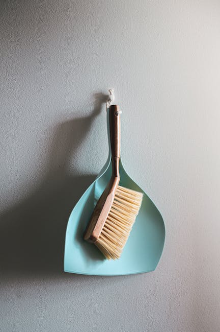 A dustpan and a brush with natural wood handles hanging on a white wall. The dustpan is made of light blue plastic, and the brush has cream-colored bristles. The tools are neatly positioned, with the broom resting inside the dustpan, illustrating a clean and organized setup suitable for surface cleaning and maintenance in domestic environments. The lighting is soft and even, emphasizing the neatness and readiness of the cleaning tools, which reflect the professional cleaning services offered by Cleaners W1, focused on providing realistic cleaning quotes in Marylebone W1 for both residential and commercial properties.