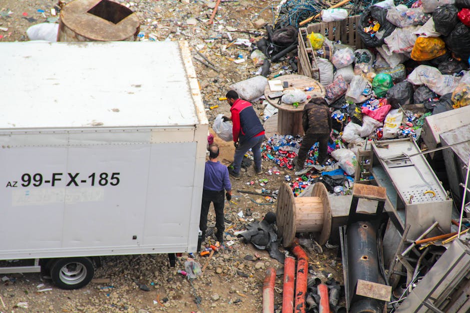 An outdoor scene depicts three individuals standing among a large, chaotic pile of mixed bulky waste, including crushed cardboard boxes, black and multicolored plastic bags, and scattered debris. The ground is covered with dirt, small rocks, and various discarded items. In the background, there are wooden spools, metal pipes, and other miscellaneous waste materials. A white truck with the registration number '99-FX-185' is partially visible to the left, parked on the uneven terrain. The individuals appear to be working or inspecting the waste, with one in a red and black jacket, another in a purple top, and the third in darker clothing. Natural lighting illuminates the scene, highlighting the mess and disarray typical of unmanaged waste disposal areas. Cleaners W1 offers professional waste management and cleaning services to ensure safe and proper disposal of bulky waste, promoting hygiene and environmental safety in residential and commercial settings.