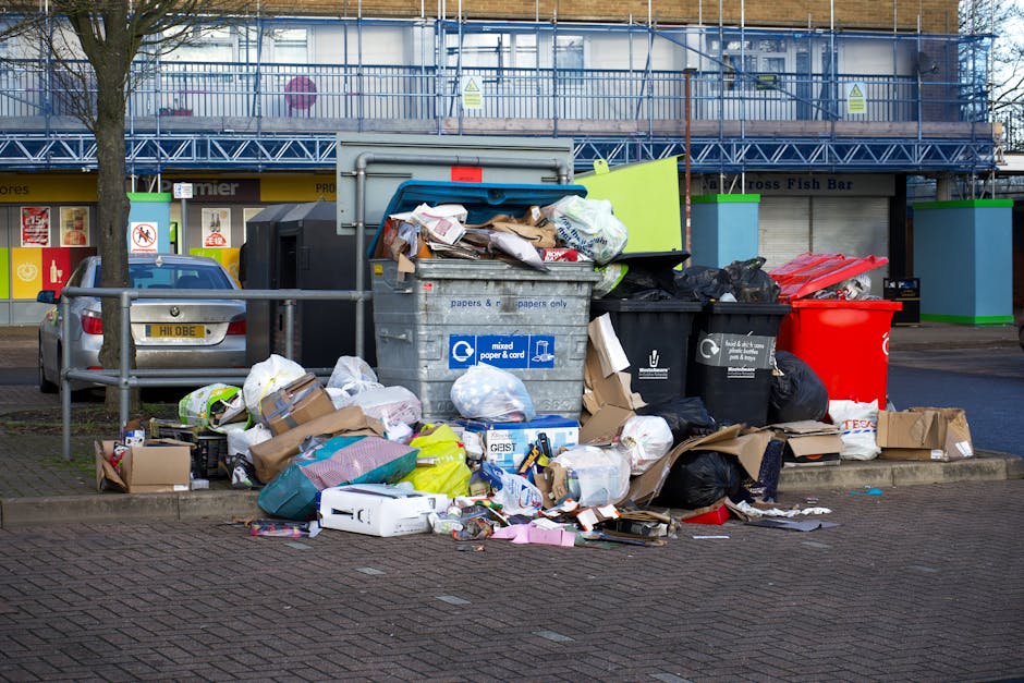 A large pile of mixed bulky waste, including cardboard boxes, paper, plastic bags, and various household items, is gathered around overflowing waste bins on a paved outdoor area. The backdrop features a building with a blue scaffolding structure, glass windows, and signage indicating commercial premises. The waste bins, some with open lids, reveal additional litter such as packaging and discarded materials. There is a metal railing separating the waste area from the road, with parked cars visible nearby. The area appears untidy and in need of proper disposal or cleaning, highlighting the importance of safe waste management and surface cleaning practices. Cleaners W1 offers professional cleaning solutions to ensure hygiene and cleanliness in residential and commercial environments, supporting effective waste disposals and deep cleaning processes.
