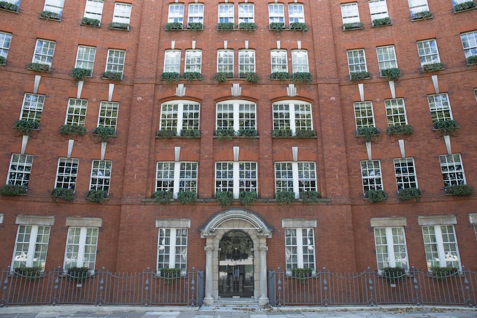 The image depicts the front facade of a multi-storey red brick residential building with numerous evenly spaced white-framed sash windows. Each window features a small ledge adorned with green potted plants, adding a touch of greenery. At the bottom of the building, there is an arched entrance with a decorative stone surround and a glass door, flanked by metal fencing. The overall appearance is clean and well-maintained, with a bright daylight setting illuminating the building's exterior. The scene emphasizes the building's classic architectural style and attention to outdoor aesthetic details, fitting for a city like Marylebone. For professional cleaning services, including surface cleaning and deep sanitisation, Cleaners W1 offers tailored solutions for property maintenance in this area.