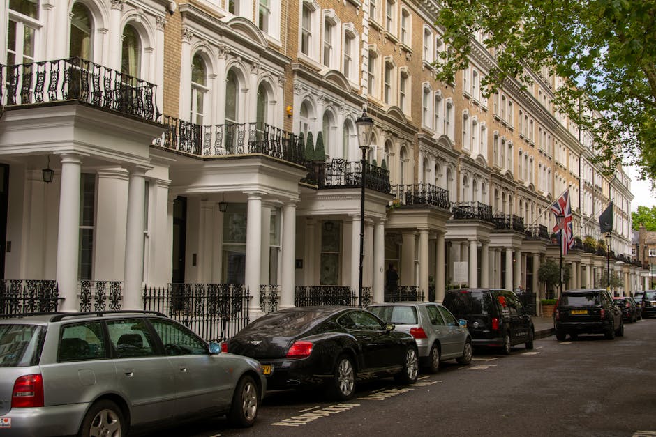 Photograph of a row of elegant Victorian-style terraced houses on a residential street in Marylebone, W1, London, featuring white facades with decorative moldings and black wrought-iron balconies. The street is lined with parked cars including a silver estate, a black coupe, and several other vehicles, with a black lamppost and British flags visible. Trees with green foliage partially shade the scene, and the overall appearance of the buildings and street is clean, well-maintained, and orderly, reflecting a typical upscale London neighborhood.