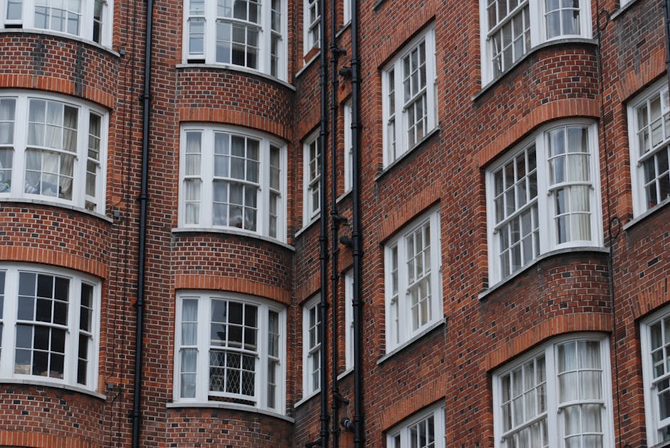 The image depicts the front facade of a multi-storey red brick residential building with numerous evenly spaced white-framed sash windows. Each window features a small ledge adorned with green potted plants, adding a touch of greenery. At the bottom of the building, there is an arched entrance with a decorative stone surround and a glass door, flanked by metal fencing. The overall appearance is clean and well-maintained, with a bright daylight setting illuminating the building's exterior. The scene emphasizes the building's classic architectural style and attention to outdoor aesthetic details, fitting for a city like Marylebone. For professional cleaning services, including surface cleaning and deep sanitisation, Cleaners W1 offers tailored solutions for property maintenance in this area.