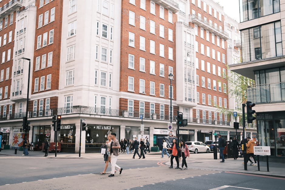 A busy street scene on Marylebone High Street, W1, showing a large multi-story building with a white and red-brick facade. The ground floor houses various retail stores with large glass windows displaying merchandise, including a café with outdoor seating. Pedestrians are crossing the street, some carrying shopping bags, while others walk along the sidewalk. The street features several traffic lights, lampposts, and parked cars, with modern buildings and greenery visible in the background. The cleanliness of the area appears well-maintained, reflecting effective surface cleaning and hygiene practices. The scene emphasizes the importance of diligent day-to-day cleaning, as promoted by Cleaners W1, for maintaining the appearance and hygiene of commercial and retail spaces.