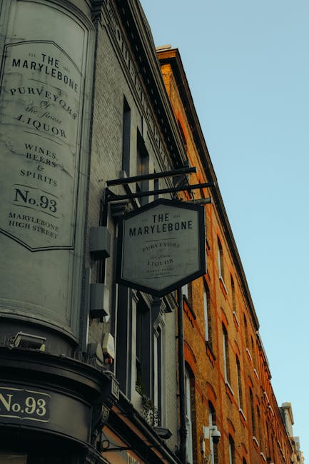 A street-level view of The Marylebone, a historic building on Marylebone High Street. The exterior features a rounded, cream-colored façade with vintage-style signage advertising liquor, wines, and spirits, including the building number 93. The signage is mounted on the wall, with one sign extending perpendicular to the building. Adjacent, there is a brick building with multiple windows, illuminated by natural daylight, contributing to a bright, clean appearance. This scene reflects the well-maintained façade characteristic of commercial storefronts in the area, emphasizing the importance of surface cleaning and maintenance to uphold the building's aesthetic appeal, in line with the day-to-day cleaning guidance from Cleaners W1 for shops on Marylebone High Street.