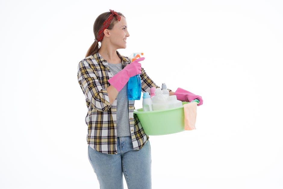 A woman holding a green plastic basin filled with various cleaning supplies, including spray bottles and cloths, dressed in casual attire with pink rubber gloves and a red checkered headband, standing next to a clean, white background. The scene depicts surface cleaning and sanitisation tasks typical of domestic or commercial cleaning services provided by Cleaners W1, emphasizing efficiency and hygiene for same-day emergency cleaning in Marylebone, W1.