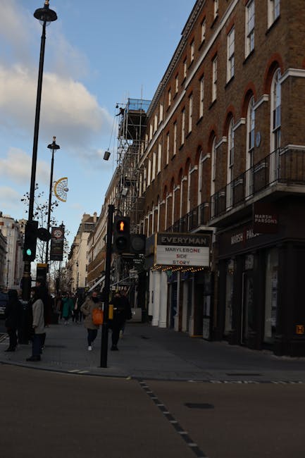 A busy street scene in Marylebone, London, featuring a row of multi-story brick buildings with large windows and white trim, some with small balconies. The street is lined with various shops, including a sign for 'Everman' and 'Marvlecore.' Pedestrians walk across the crosswalk under streetlights, with some carrying shopping bags. Traffic lights are visible, showing a red pedestrian signal. The sky is partly cloudy, and scaffolding is present on one building, indicating ongoing maintenance. The area appears clean and well-maintained, with a focus on pedestrian activity and urban street furniture, illustrating typical street-level commercial and residential environment in Westminster.