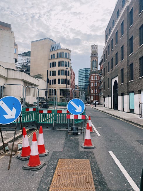 A city street in Marylebone during daytime, featuring construction barriers with blue and white directional signs, orange traffic cones, and a metal manhole cover in the foreground. Surrounded by modern and historic buildings with various window styles, the street is partially blocked off for street works, with no visible pedestrians or vehicles. Overcast sky creates diffused lighting, and the scene emphasizes urban maintenance and safety protocols typical of street clearance activities. Cleaners W1 specializes in surface cleaning and deep cleaning, ensuring hygienic and well-maintained commercial and residential environments, as part of compliance with local street clearance rules in W1.