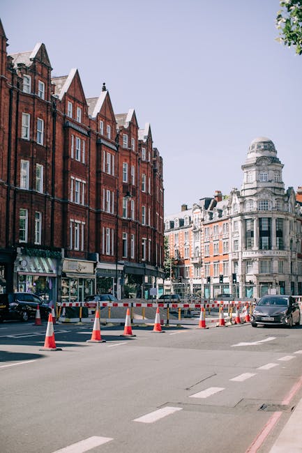 A busy street scene in Marylebone, London, featuring a row of multi-story brick buildings with large windows and white trim, some with small balconies. The street is lined with various shops, including a sign for 'Everman' and 'Marvlecore.' Pedestrians walk across the crosswalk under streetlights, with some carrying shopping bags. Traffic lights are visible, showing a red pedestrian signal. The sky is partly cloudy, and scaffolding is present on one building, indicating ongoing maintenance. The area appears clean and well-maintained, with a focus on pedestrian activity and urban street furniture, illustrating typical street-level commercial and residential environment in Westminster.
