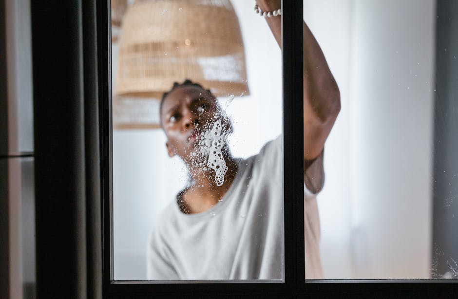A woman is seen cleaning a glass window from the inside of a home or apartment, using a cloth or sponge to wipe away soap suds and water. The window has visible foam and streaks, indicating active surface cleaning and sanitisation. The scene is illuminated by natural daylight, which highlights the transparency and cleanliness of the glass surface. Behind the window, the interior of the room features a woven lampshade hanging from the ceiling and a neutral-colored wall, suggesting a modern, well-maintained living space. This image reflects detailed surface cleaning and hygiene practices typical of domestic cleaning, as performed by professionals like Cleaners W1 in Marylebone, as promoted on the webpage about transparent pricing and avoiding hidden cleaning fees.