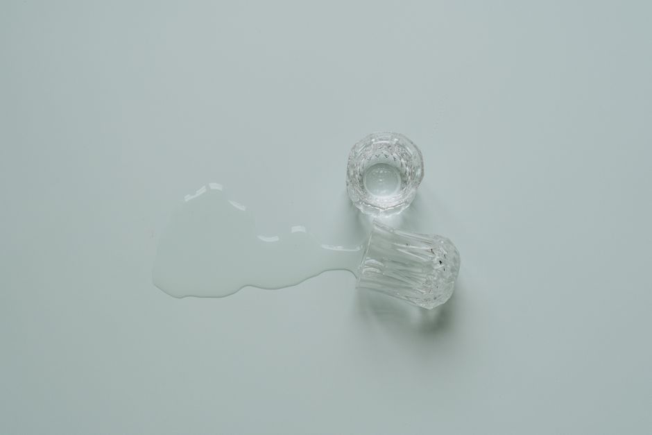 A clear glass tumbler and a small glass jar with faceted sides are placed on a smooth, white surface. The tumbler is lying on its side with some water spilled around it, creating a reflective, wet patch on the surface. The jar is positioned upright nearby, with its open top visible. The overall scene appears clean and minimalistic, highlighting surface cleaning and sanitisation practices typical of domestic and commercial environments. The lighting is bright and even, emphasizing the clarity and cleanliness of the glass and the surface. This image is used to illustrate transparent pricing and clear service descriptions by Cleaners W1 on their Marylebone cleaning services webpage.