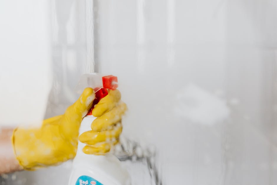 A woman is seen cleaning a glass window from the inside of a home or apartment, using a cloth or sponge to wipe away soap suds and water. The window has visible foam and streaks, indicating active surface cleaning and sanitisation. The scene is illuminated by natural daylight, which highlights the transparency and cleanliness of the glass surface. Behind the window, the interior of the room features a woven lampshade hanging from the ceiling and a neutral-colored wall, suggesting a modern, well-maintained living space. This image reflects detailed surface cleaning and hygiene practices typical of domestic cleaning, as performed by professionals like Cleaners W1 in Marylebone, as promoted on the webpage about transparent pricing and avoiding hidden cleaning fees.