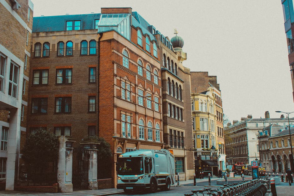 A city street scene in Marylebone W1, featuring multi-storey buildings with a mix of brick and pale yellow facades, large windows, and decorative architectural details. In the foreground, a blue waste collection truck labeled 'Dennis' is parked on the cobblestone street. Surrounding it are metal bollards, parked bicycles, and a lamppost, with additional vehicles and historic buildings visible in the background. The street appears clean, with no visible litter or debris, and the daylight provides natural lighting that highlights the textured surfaces of the brick and stone buildings. This scene reflects an urban environment where professional cleaning services, such as those provided by Cleaners W1, could be essential for maintaining hygiene and compliance with waste disposal regulations relevant to Westminster Council fines for commercial waste in Marylebone W1.