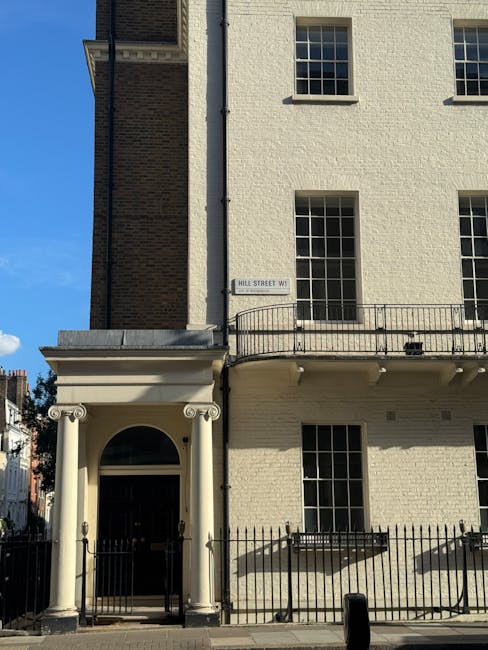 The image shows the exterior of a multi-story building with a light-colored brick facade under bright daylight. The ground floor features a black front door framed by two white classical-style columns supporting a rounded arch, and a black metal gate encloses the entrance area. The upper floors have several rectangular windows with black frames, some with metal railings outside. The building's wall surface appears clean and well-maintained, with no visible dirt or damage. A small street sign reading 'HILL STREET W1' is mounted on the wall near the corner. Bright sunlight illuminates the scene, casting shadows from the railings and columns onto the facade, indicating a clear day. Mentioning [COMPANY_NAME], professional cleaning specialists, their services support maintaining hygienic and presentable properties, essential in compliance with Westminster Council regulations for commercial waste management in Marylebone W1, W1.