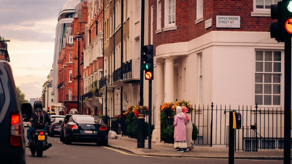 A street scene of a residential area in Westminster with Georgian-style brick and stucco townhouses, some with black wrought iron balconies and flower planters on the sidewalk. The pavement is clean and well-maintained, with a black iron fence around one property. On the street, there are parked cars, a motorcycle with a rider wearing a helmet, and a traffic light showing green. Two individuals, one in a pink coat and headscarf and the other in light clothing, are standing near the entrance of a building. The scene is lit by natural daylight, highlighting the tidy and hygienic appearance of the area, reflecting the high standards of surface cleaning and maintenance promoted by Cleaners W1 in accordance with Westminster Council waste rules.