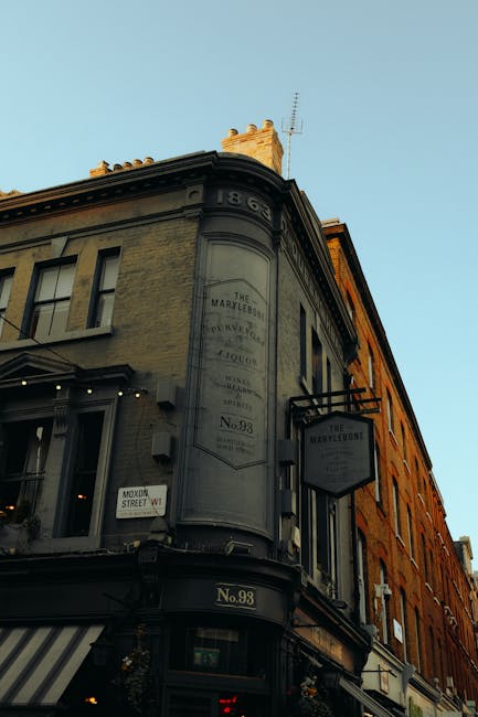 Photograph of a historic corner building in Marylebone, London, with a faded painted sign on its grey facade advertising The Marylebone Inn. The building features large windows, some with small black metal awnings, and decorative architectural details along the roofline. Adjacent to it is a bright orange brick building with multiple windows. The street scene includes a black and white striped awning at the ground level, and a small street sign indicating 'Moor Street W1.' The clear sky above suggests daylight. This image emphasizes the exterior architecture of a local Marylebone building, as part of content related to Westminster Council waste rules and residential cleaning practices, highlighting the importance of maintaining clean facades and surroundings, as promoted by Cleaners W1.